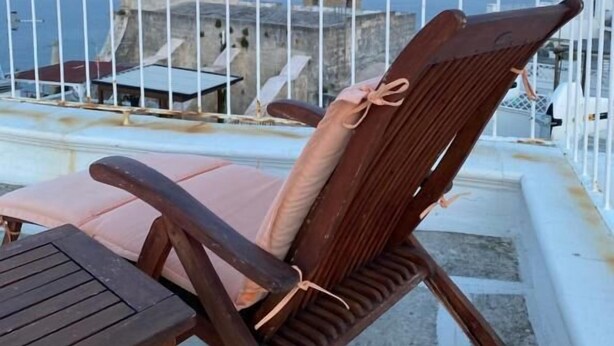 A wooden chair with a cushion and a small table on a balcony with a white railing, overlooking a coastal area with buildings and a clear sky.