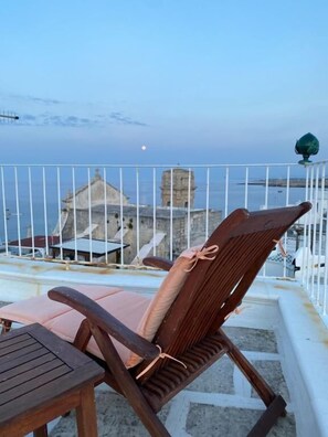 Uma cadeira de madeira com almofada e uma pequena mesa numa varanda com corrimão branco, com vista para uma área costeira com edifícios e céu limpo.