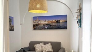 A living room with a sofa, a small table, a hanging lamp, and a painting of a cityscape.