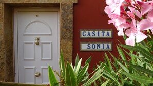 En hvid dør med messinggreb, en rød væg med to skilte der står "CASITA" og "SONIA", samt grøn vegetation i forgrunden.