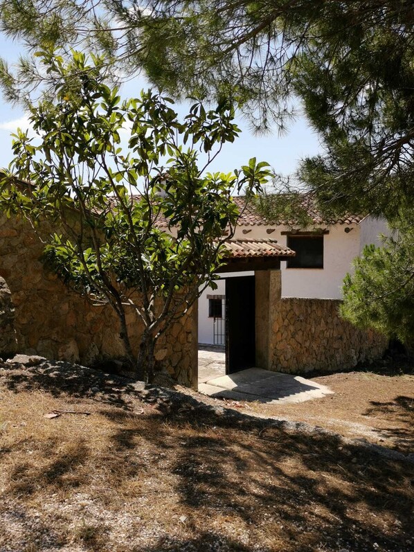 Un bâtiment en pierre avec un toit en tuiles, une porte d’entrée et un petit jardin où l’herbe est sèche et où se trouve un arbre.