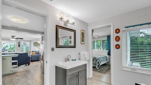 A modern, well-lit room with a large window, a bed, a bathroom with a sink and mirror, and a dining area with a table and chairs. - Mermaid Tale 2 Bedroom Condo by RedAwning (Bradenton Beach)