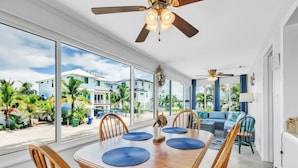 A dining area with a large table and chairs, large windows with a view of buildings and palm trees, and ceiling fans. - Mermaid Tale 2 Bedroom Condo by RedAwning (Bradenton Beach)