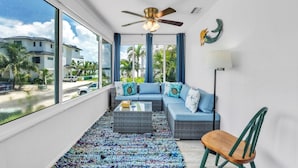 A modern living room with a large window, a ceiling fan, a sofa set, a coffee table, and a view of a residential area. - Mermaid Tale 2 Bedroom Condo by RedAwning (Bradenton Beach)