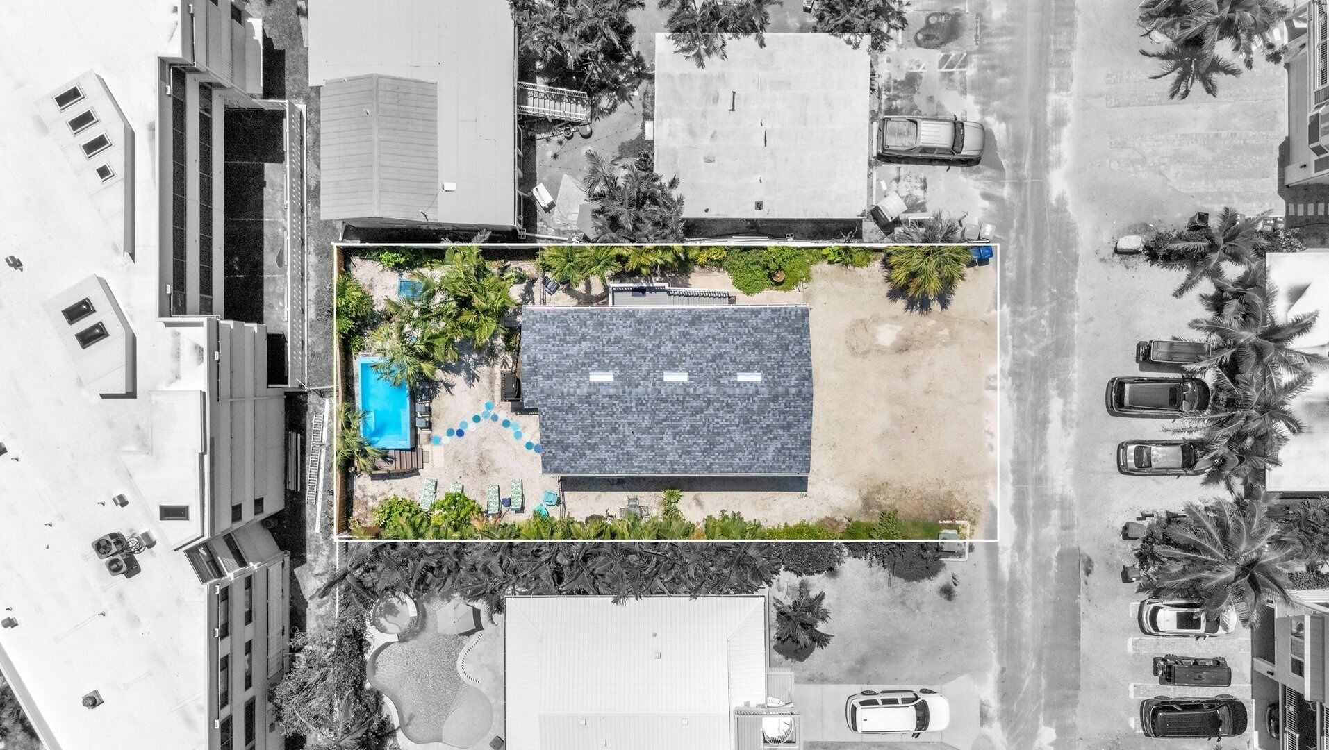 An aerial view of a modern building with a rooftop pool, surrounded by palm trees and adjacent parking areas.
