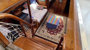 Interior de barco com banco de madeira, mesa pequena, assento acolchoado e tapete estampado.