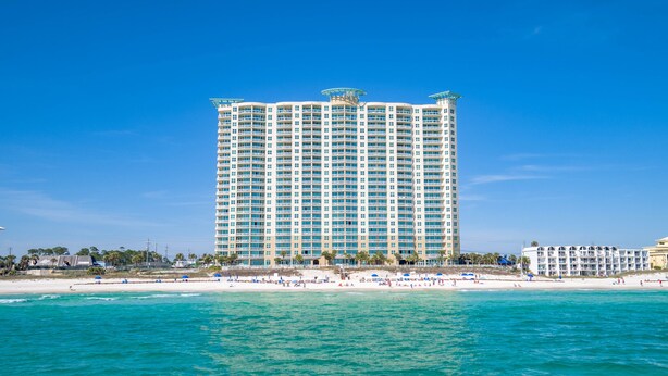 A tall hotel with multiple balconies, located near a beach with clear blue waters.