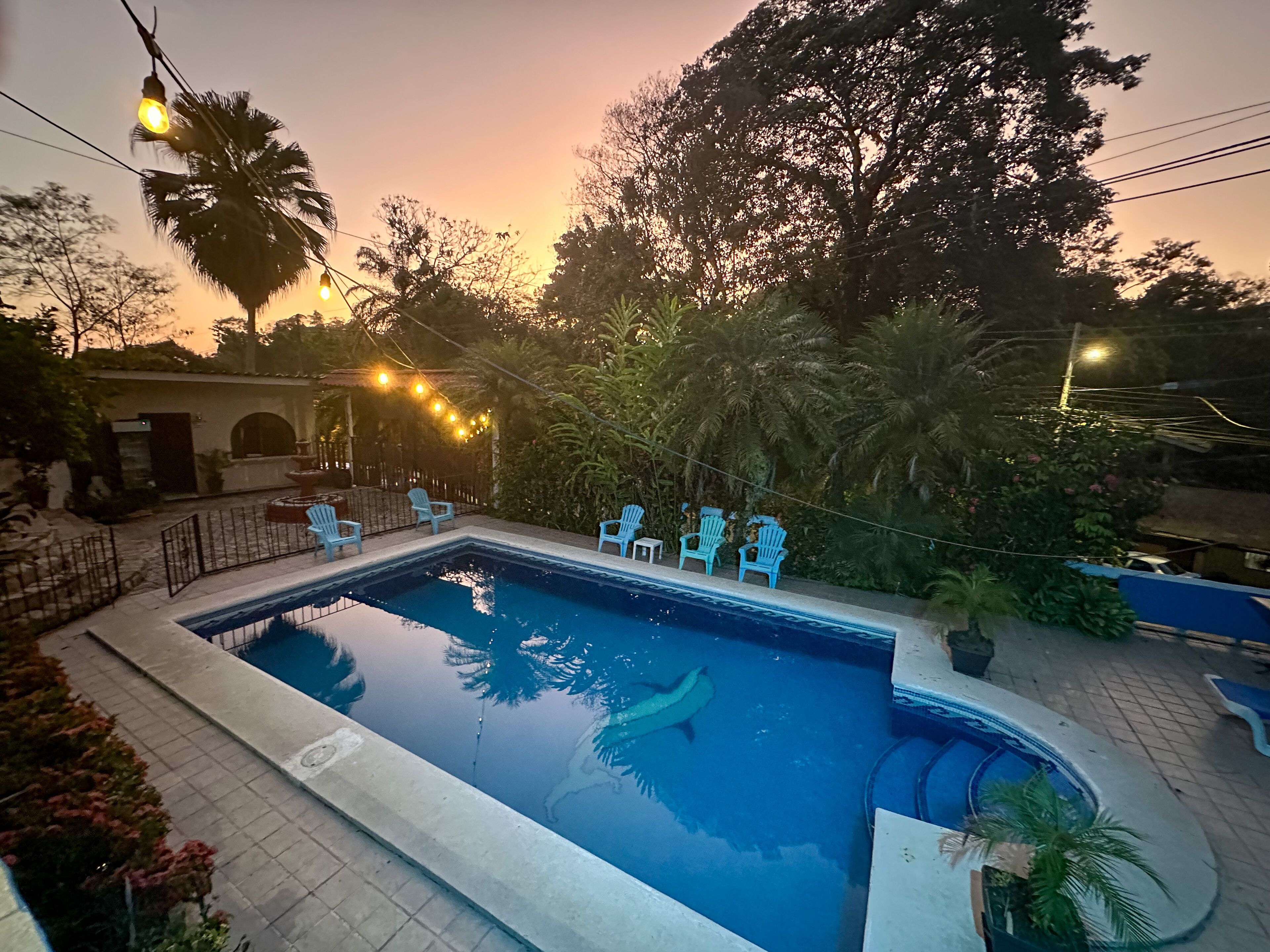 Una piscina con superficie de agua azul clara, rodeada por una solana con baldosas. Varias sillas azul claro están dispuestas alrededor de la piscina, y una guirnalda de luces es visible al fondo.