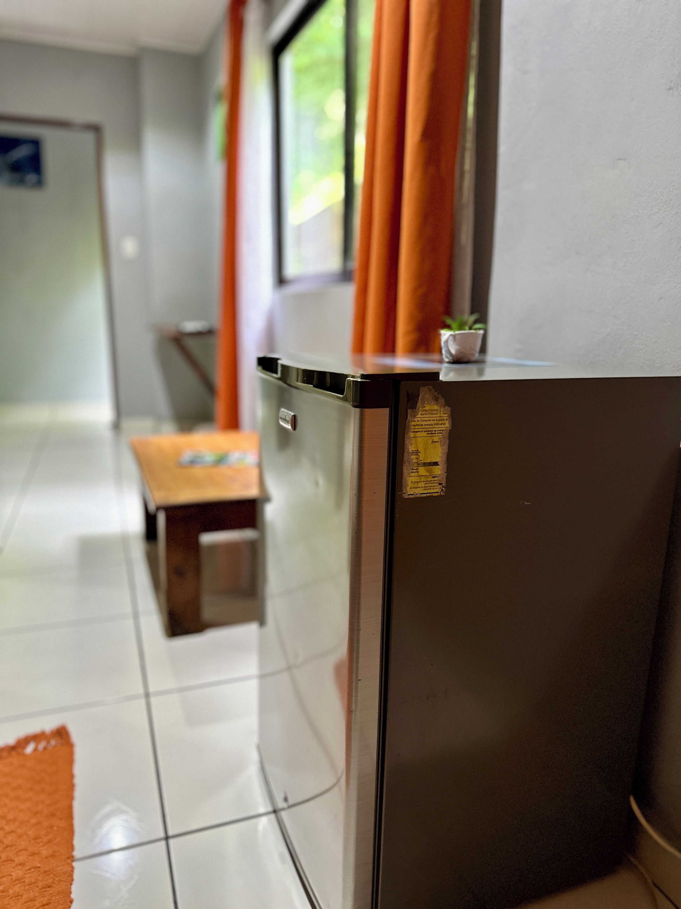 A compact kitchen area with a refrigerator, a small table, and a window with curtains.