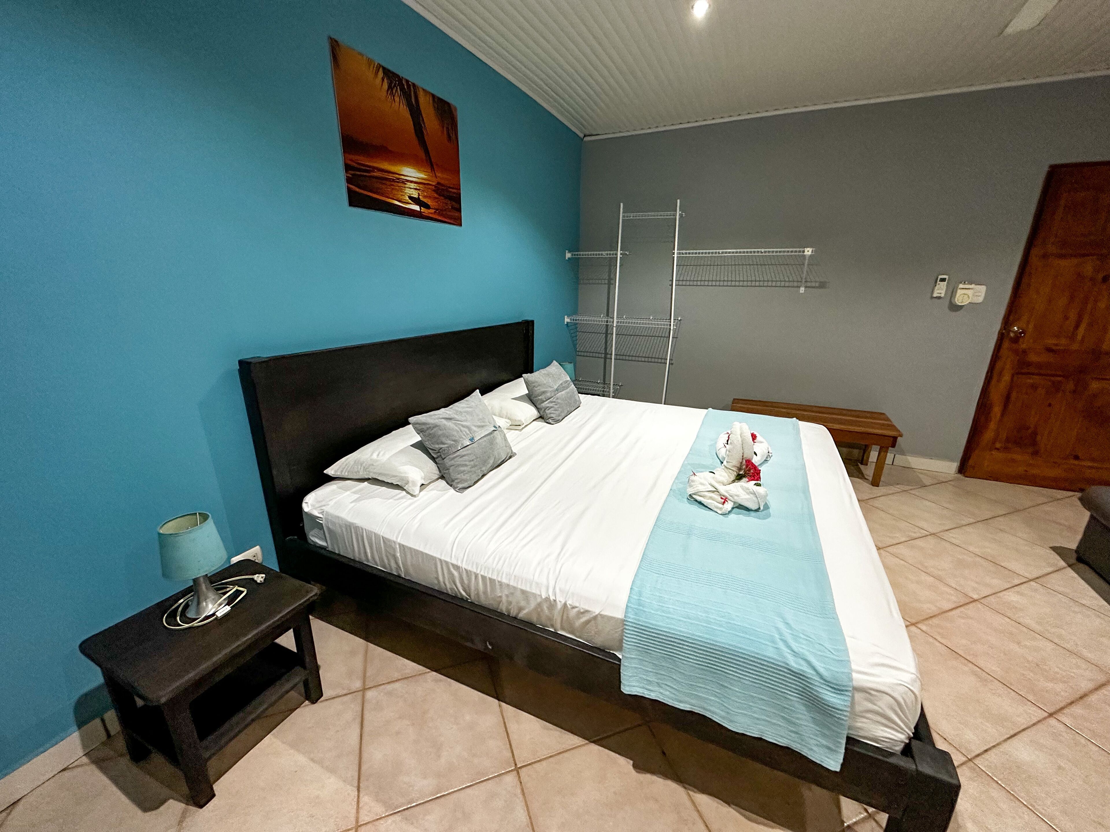 A neatly made bed with pillows, a bedside table with a lamp, a wall-mounted drying rack, and a wooden bench.