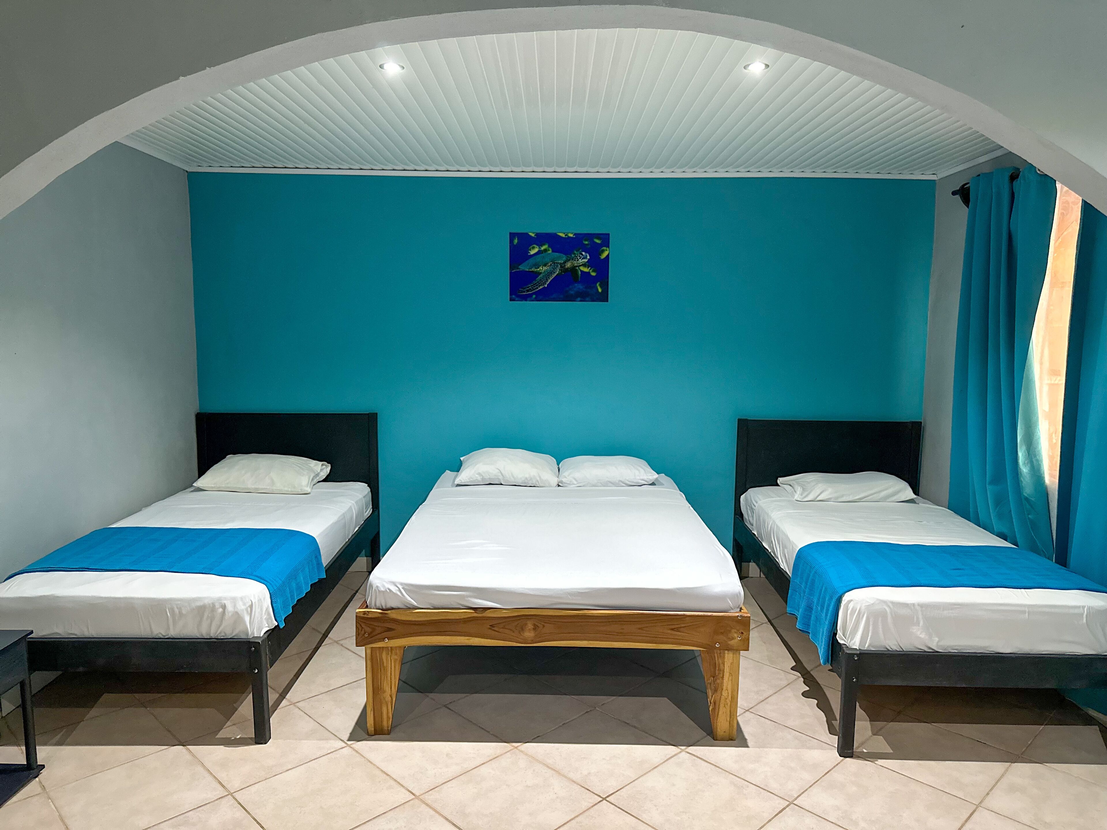Una habitación con dos camas individuales y una cama más grande, todas con ropa blanca. Las paredes están pintadas de verde azulado y el techo tiene un diseño acanalado.