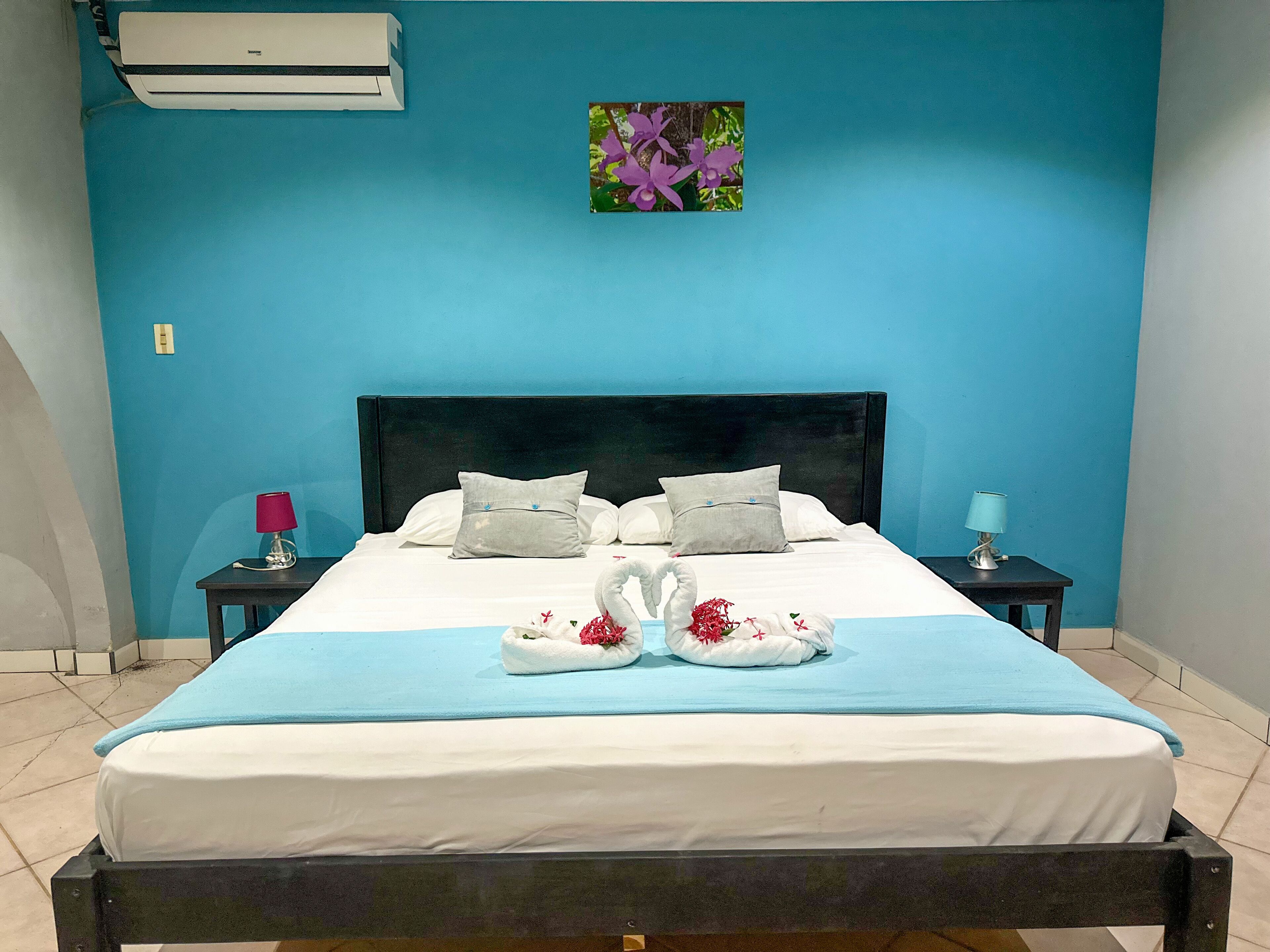 Une chambre d’hôtel comprenant un lit, une table de chevet, une lampe, un climatiseur et un tableau accroché au mur.