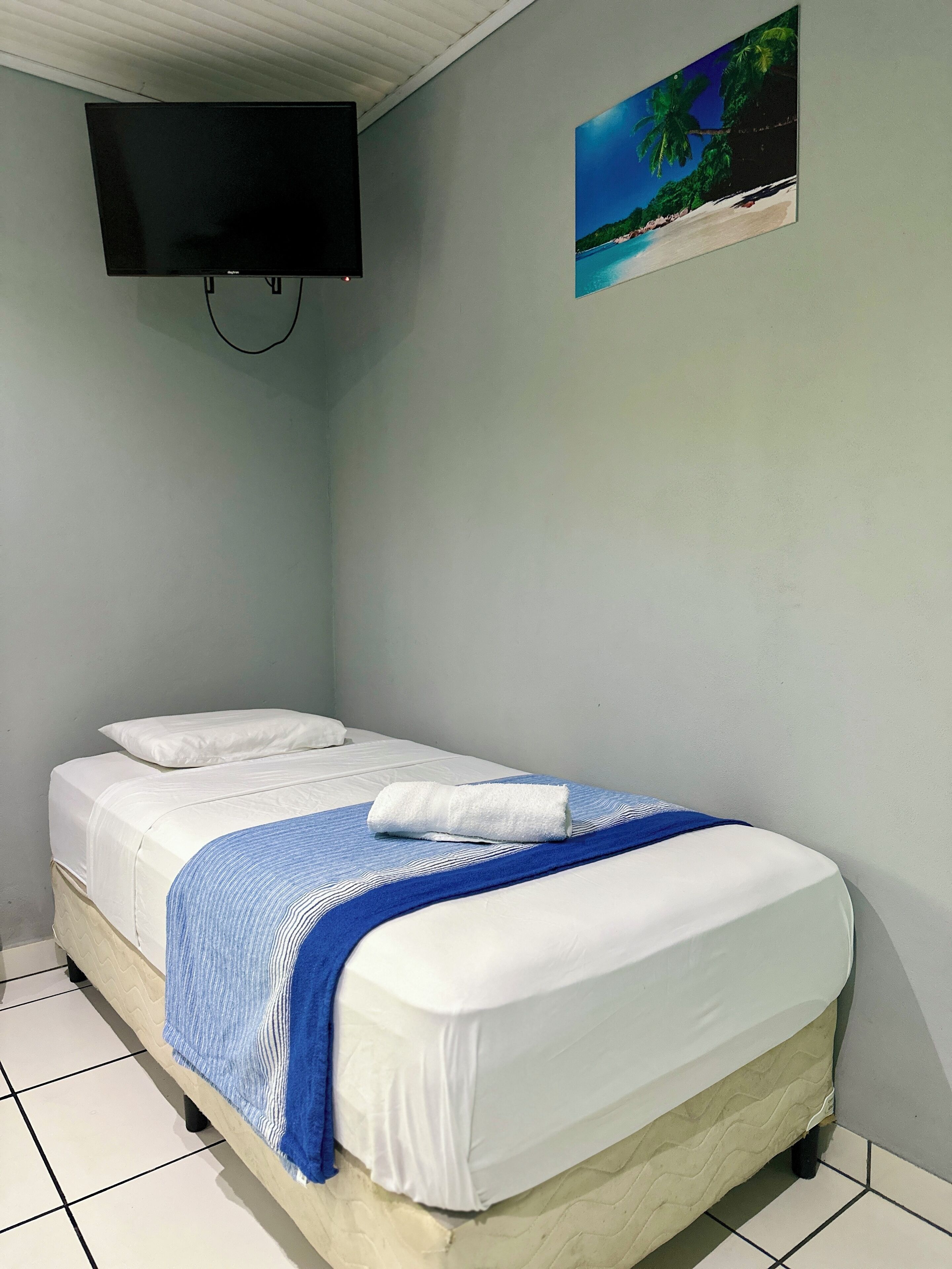 Un lit simple avec une literie blanche et une couverture à rayures bleues, un téléviseur à écran plat fixé au mur et un tableau représentant une plage tropicale au-dessus du lit.