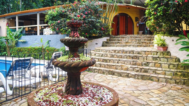 A stone staircase leading to a building with a red door, surrounded by lush greenery and a decorative fountain. - Hotel Colibri (Quepos)