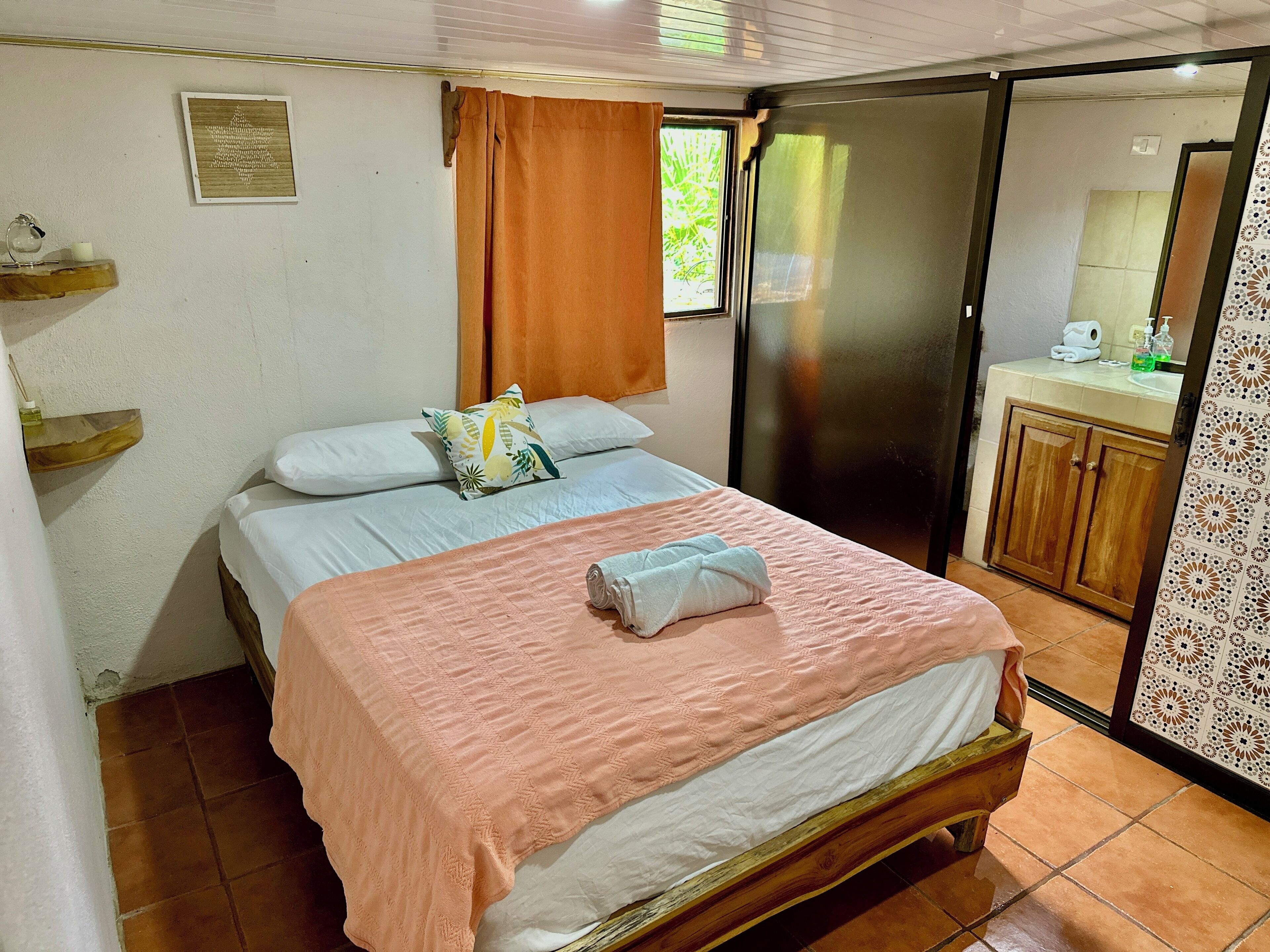 A bedroom with a bed, a wooden headboard, a window with curtains, a bathroom with a sink and mirror, and a door leading to another room.