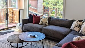 Un salon moderne avec un canapé d’angle, une table basse ronde et de grandes fenêtres donnant sur un jardin et les bâtiments voisins.