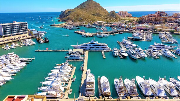 Un port de plaisance où sont amarrés de nombreux yachts, un grand bâtiment sur la gauche et une colline imposante en arrière-plan.