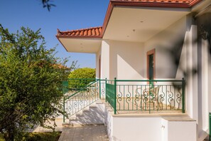 Un bâtiment à l’extérieur blanc, avec un toit recouvert de tuiles rouges, un garde-corps métallique vert et un escalier menant à un espace abrité où se trouve un banc.
