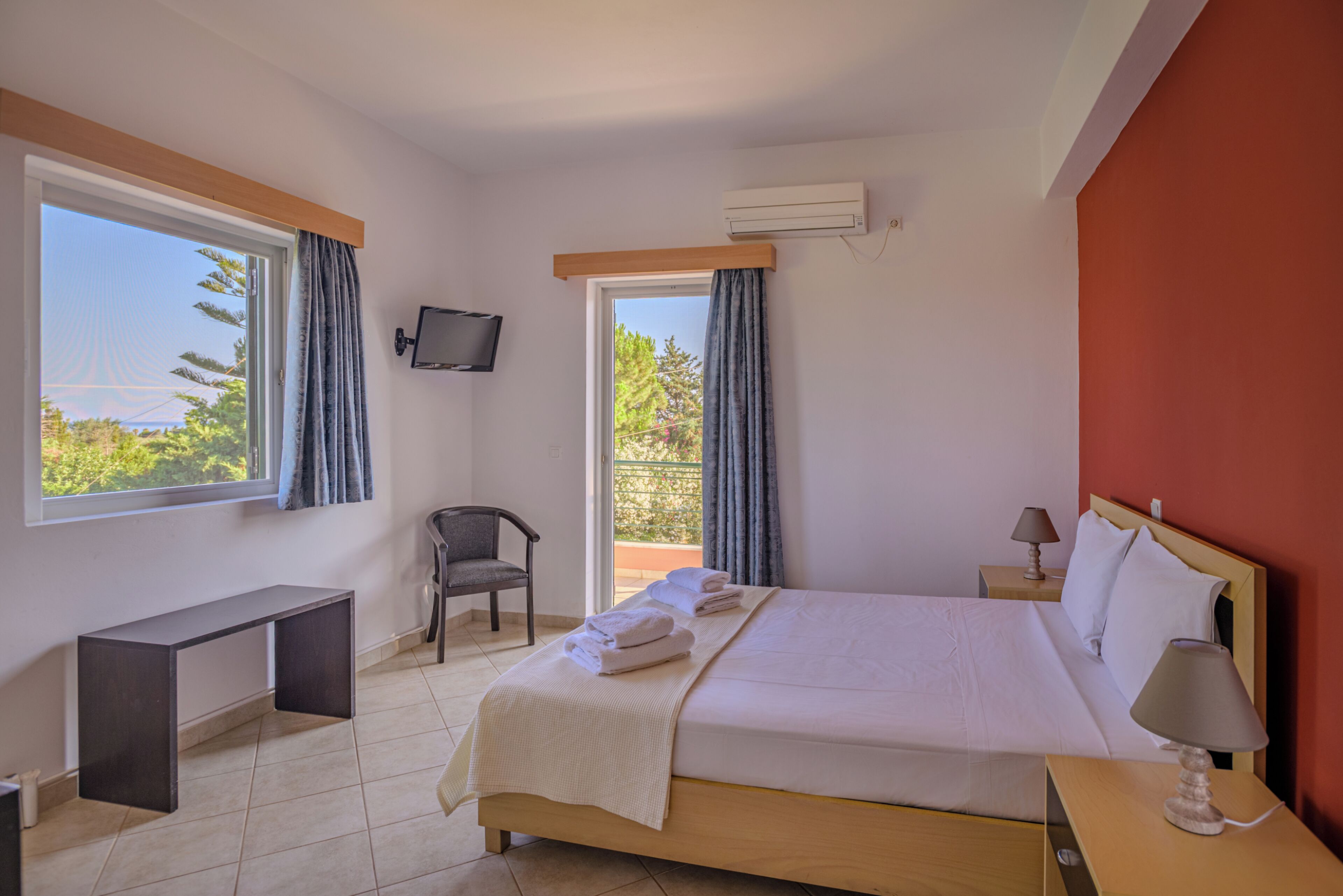 A hotel room with a bed, a desk, a chair, a TV, and a balcony with curtains.