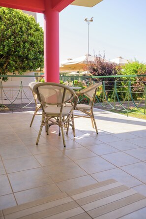 A covered outdoor seating area with two wicker chairs and a table, surrounded by a green fence and a red pillar. - Stomio Villague (Trifylia)