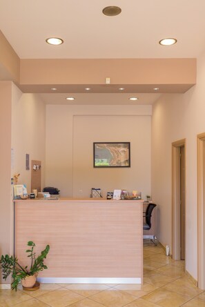 A reception desk with a potted plant, a framed picture on the wall, and a door to the right. - Stomio Villague (Trifylia)