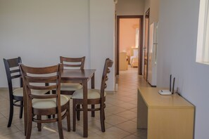 A dining area with a wooden table and chairs, a small kitchenette with a refrigerator, and a hallway leading to a bedroom. - Stomio Villague (Trifylia)