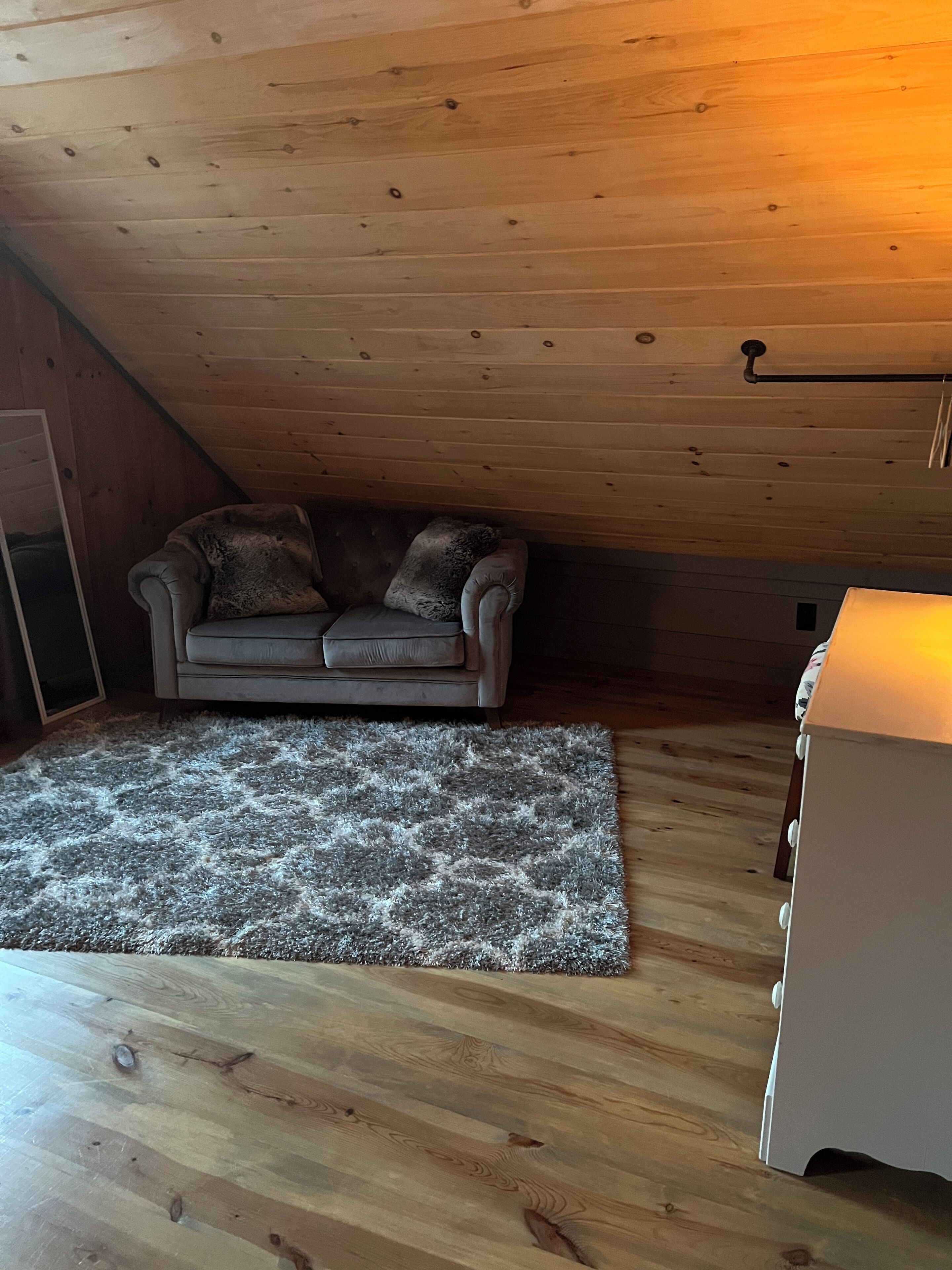 A cozy attic room with a wooden ceiling, a grey sofa with two pillows, a fluffy rug, and a white cabinet.