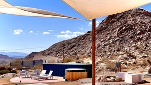 An outdoor patio with a sofa, a table, and a barbecue grill. There is a large shade structure, a small building, and a mountainous backdrop. - Stars on the Rocks: Pool/HotTub/FirePit/Boulders! (Joshua Tree)