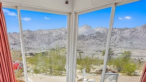 A bedroom with a bed, a desk, and a chair. It has large windows with a view of a rocky landscape. - Stars on the Rocks: Pool/HotTub/FirePit/Boulders! (Joshua Tree)