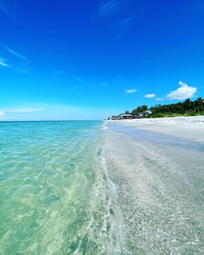 Una playa con agua turquesa cristalina, arena blanca y algunos edificios al fondo.