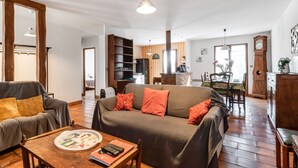 Un salon spacieux comprenant un canapé, une table basse avec un livre et une assiette, un coin repas avec une table et des chaises, et un coin cuisine avec une horloge et un meuble.
