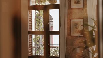 A cozy room with a wooden table, books, a glass jar, and a wicker chair. A window with a metal gate and curtains is visible, along with framed pictures on the wall.