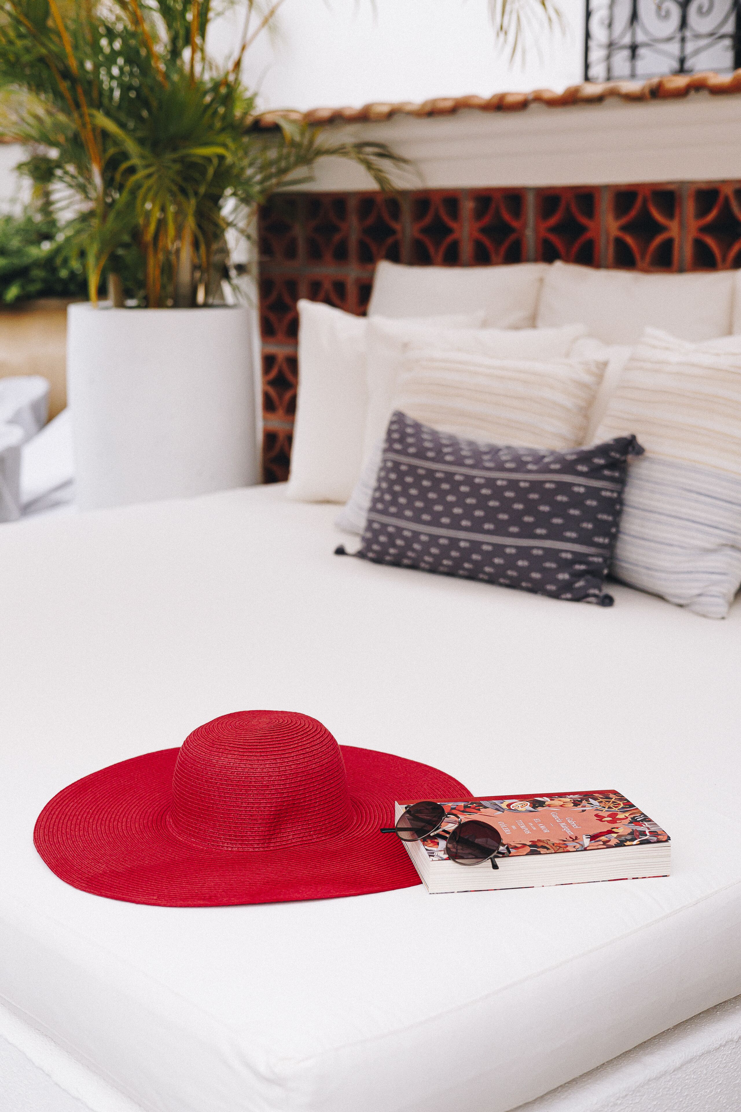 A bed with white bedding, a red hat, and a book with sunglasses on it.