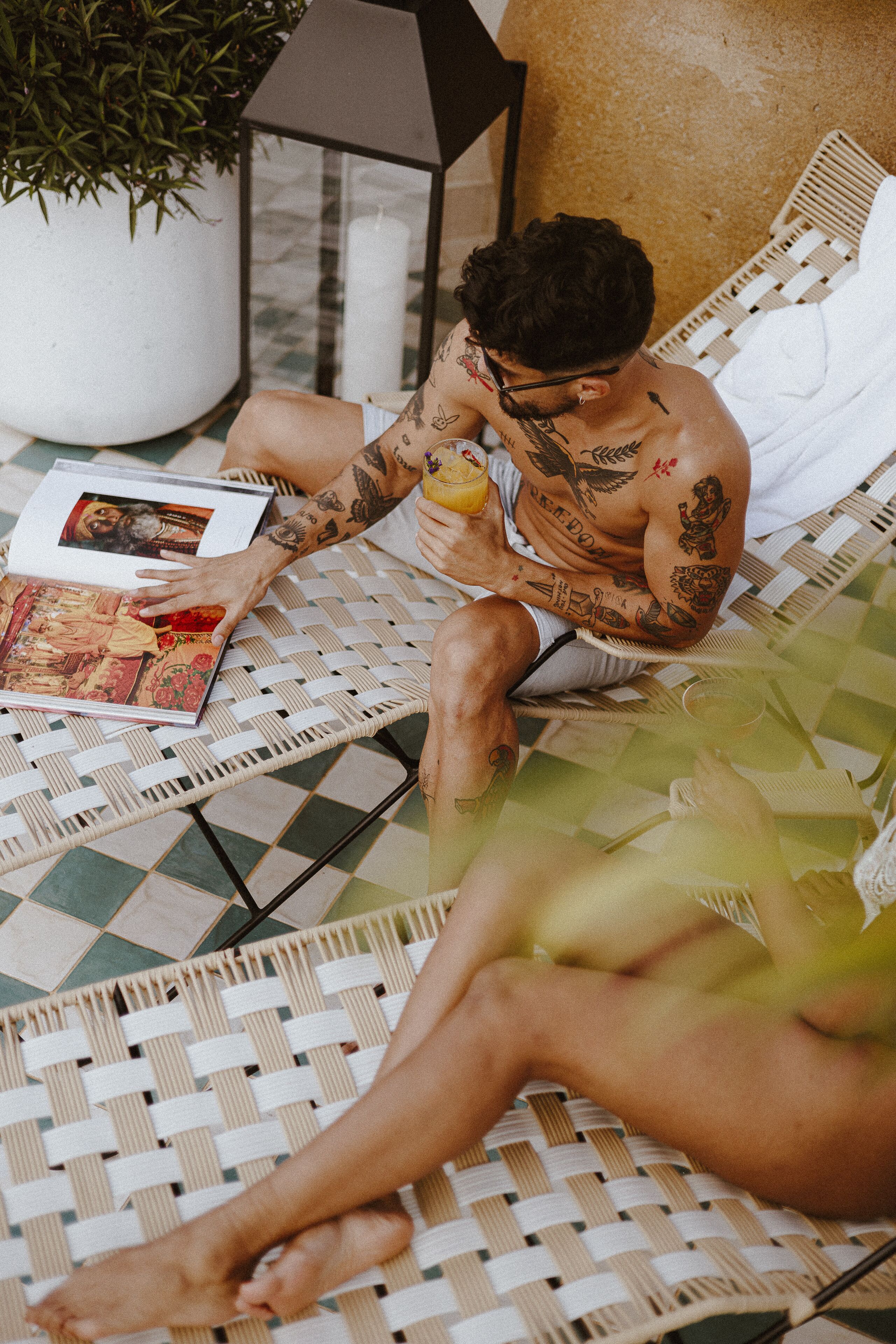 Un homme tatoué est assis sur un fauteuil en osier, lisant un magazine. On aperçoit, en arrière-plan, une plante en pot et une lampe.