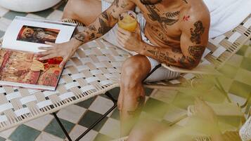 Un homme tatoué est assis sur un fauteuil en osier, lisant un magazine. On aperçoit, en arrière-plan, une plante en pot et une lampe.