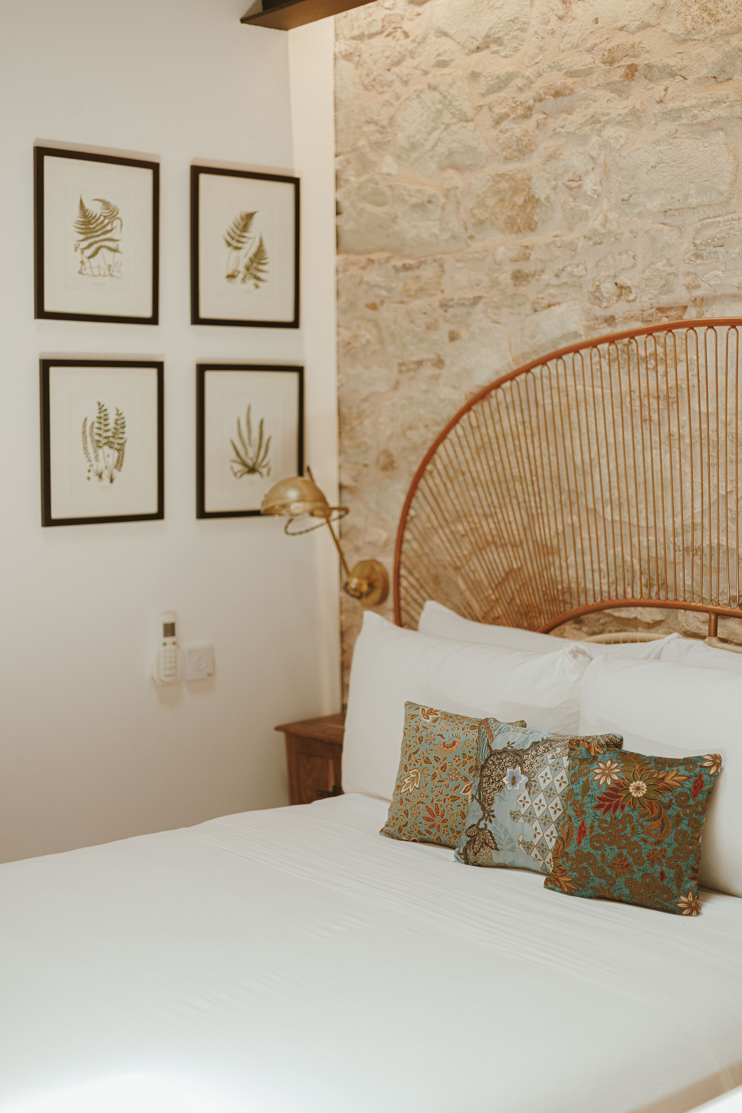 Un dormitorio con una cama, un teléfono en la pared, láminas enmarcadas con motivos botánicos y una pared de piedra texturizada.