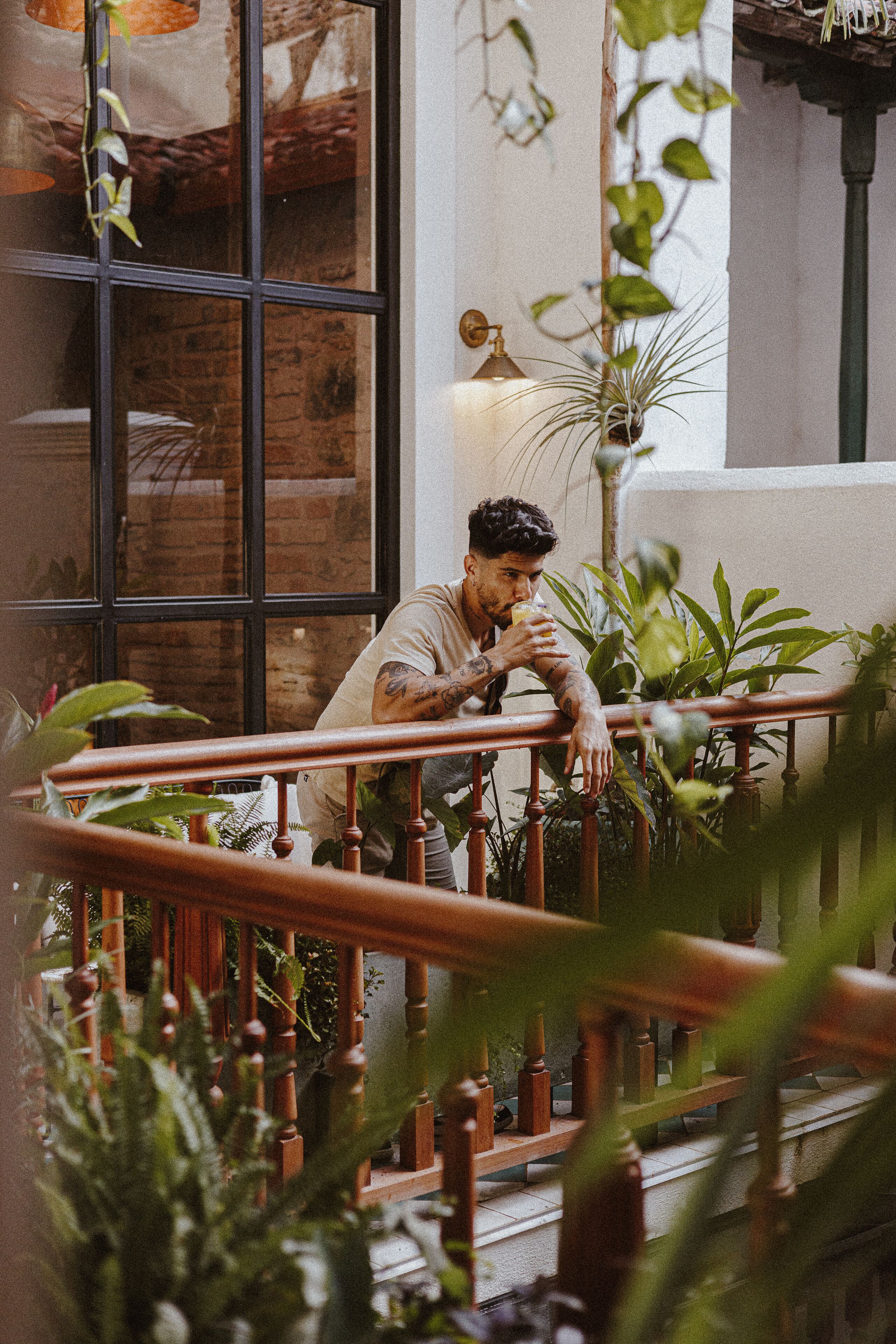 A person sitting on a wooden bench with a railing, surrounded by greenery and a brick wall.