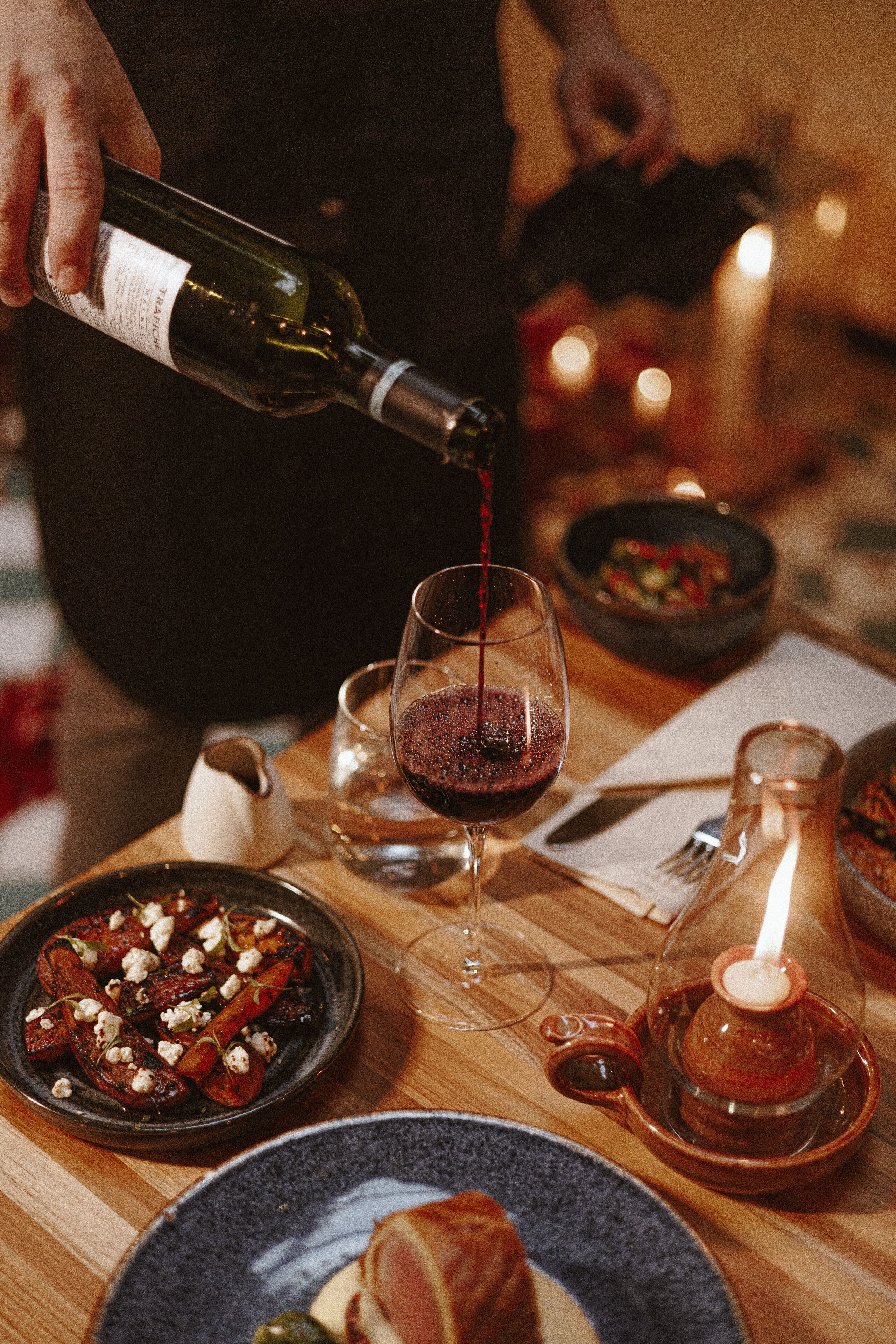 Una mesa de comedor con una botella de vino sirviéndose en una copa, acompañada de diversos platos y una vela.