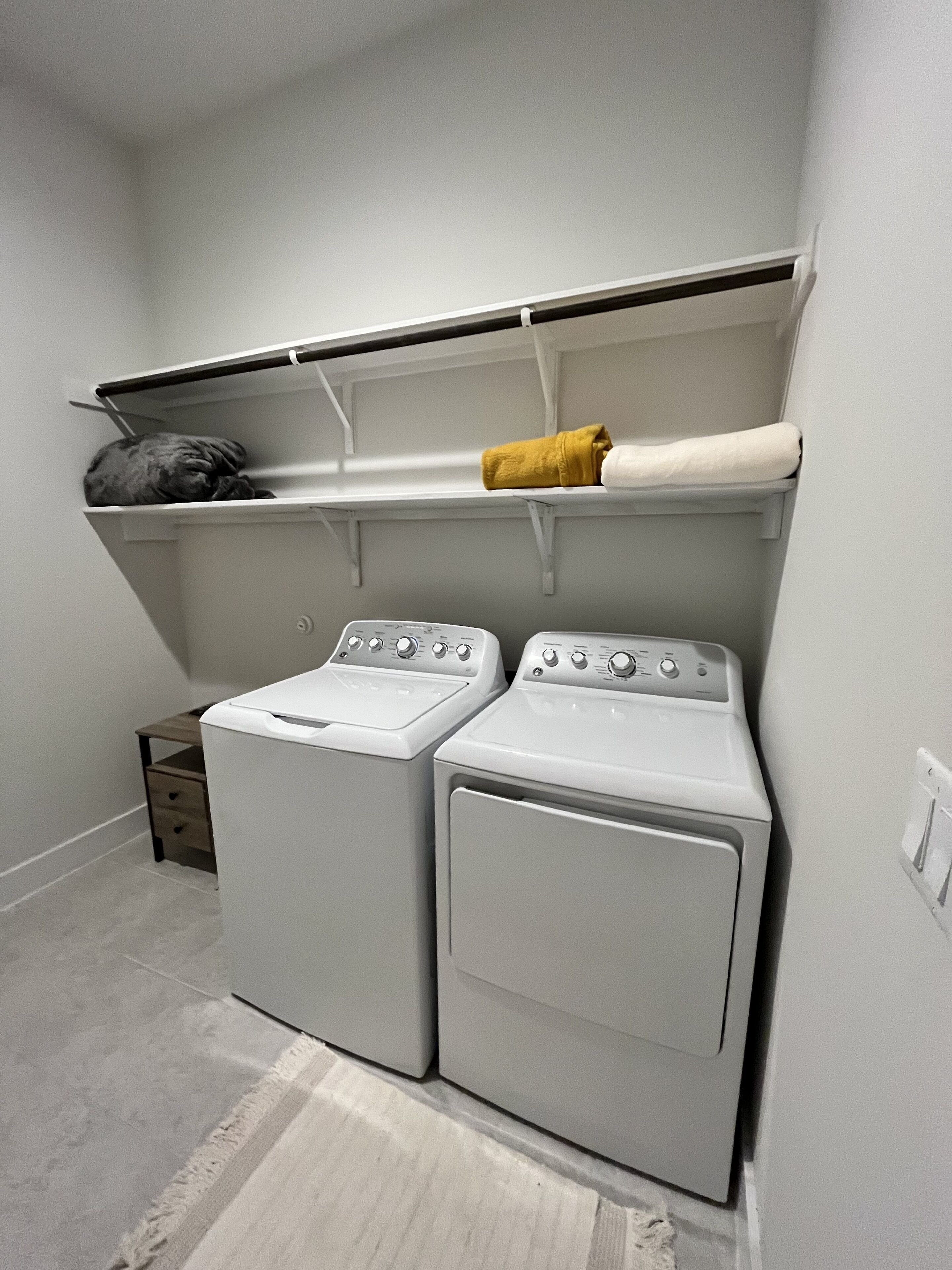 Een wasserette met twee witte wasmachines, een houten plank erboven en een klein houten kastje met lades.