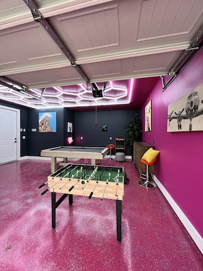 A room with a foosball table, a ceiling-mounted projector, and a wall with two paintings.