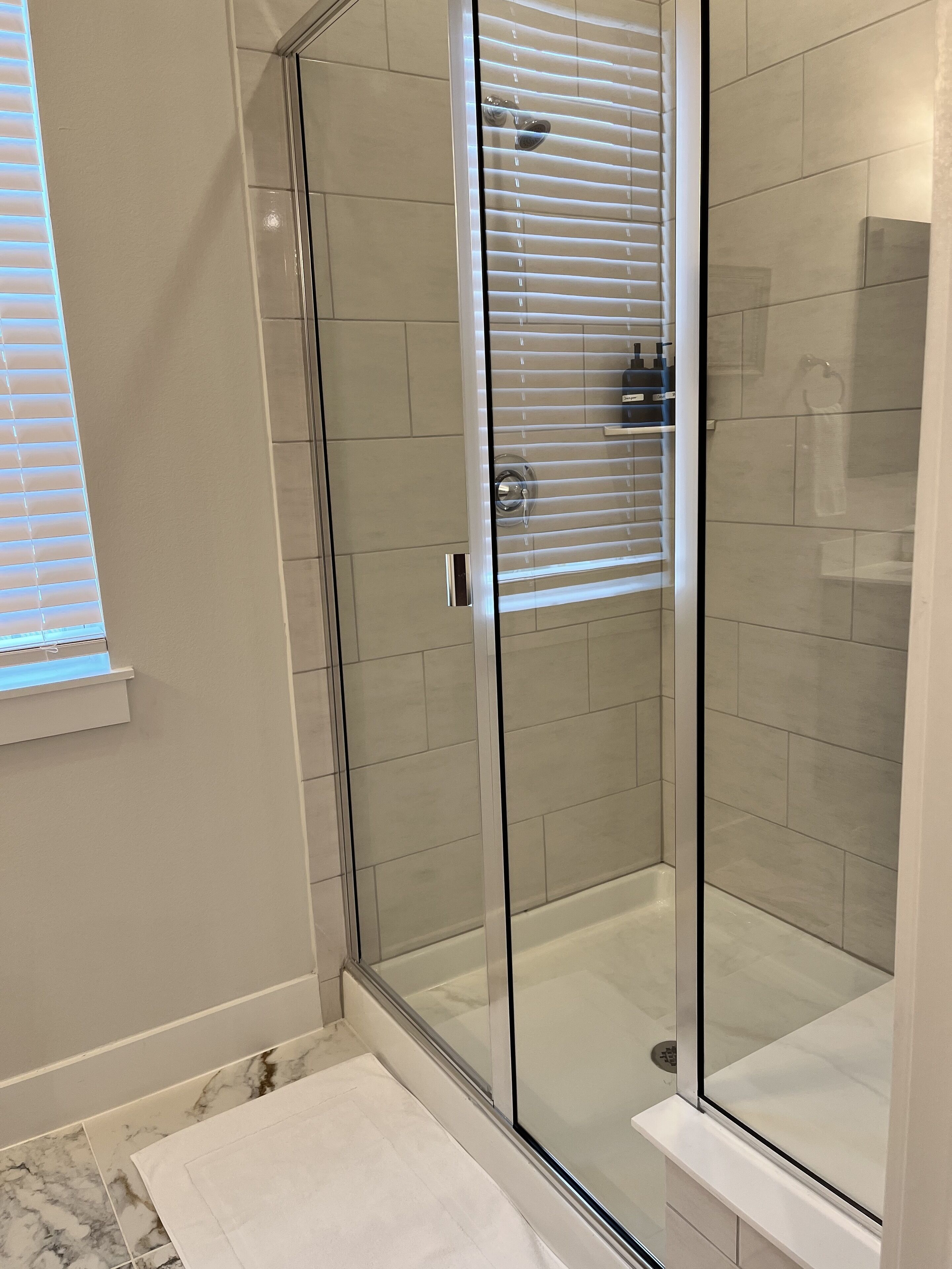 A modern bathroom with a glass-enclosed shower, white tiled walls, and a window with blinds.