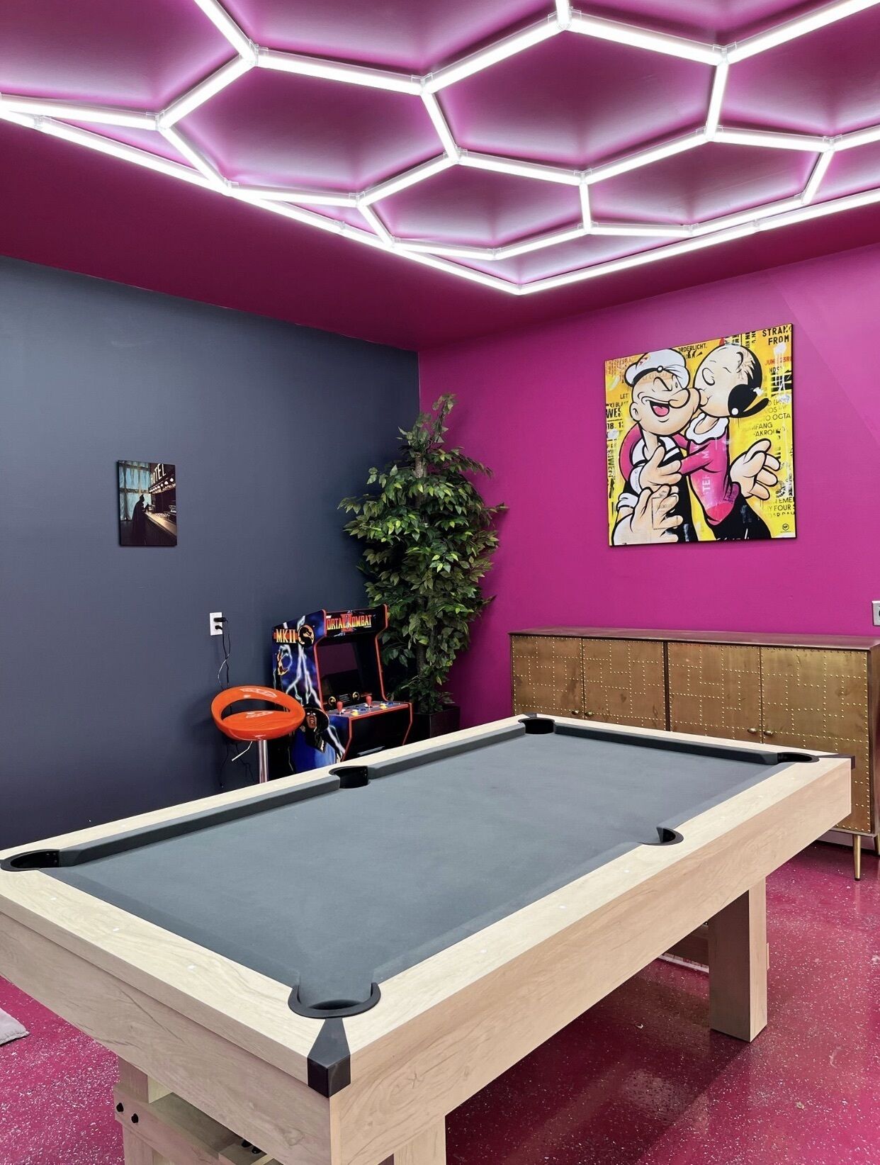 A pool table in a room with a vibrant pink and blue color scheme, a large green plant, and a retro arcade machine.