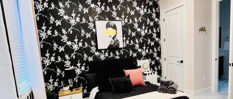 A bedroom with a black and white floral wallpaper, a chandelier, a bed with black and pink bedding, and a round rug with ottomans.