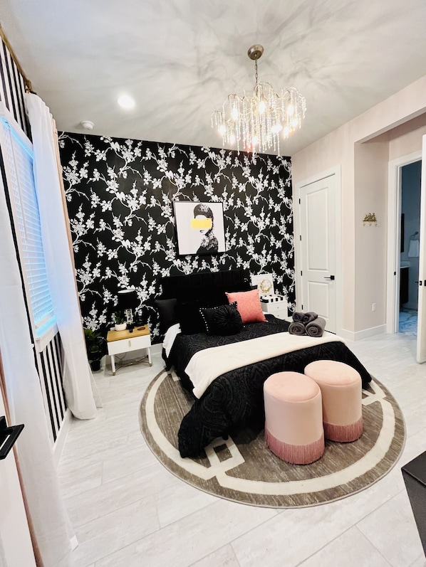 A bedroom with a black and white floral wallpaper, a chandelier, a bed with black and pink bedding, and a round rug with ottomans.