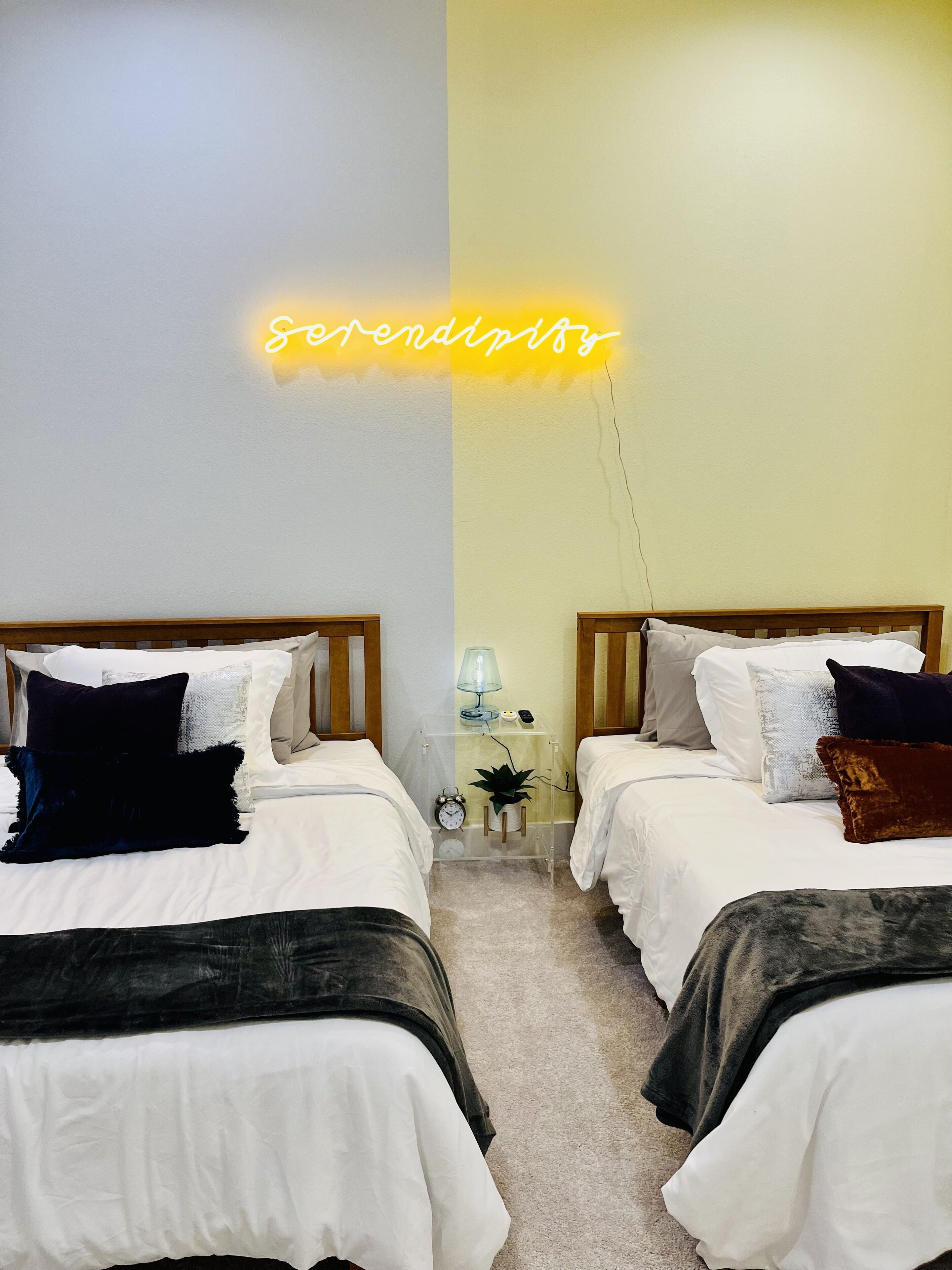 A bedroom with two beds, a nightstand with a lamp, a clock, and a neon sign reading "serenity".