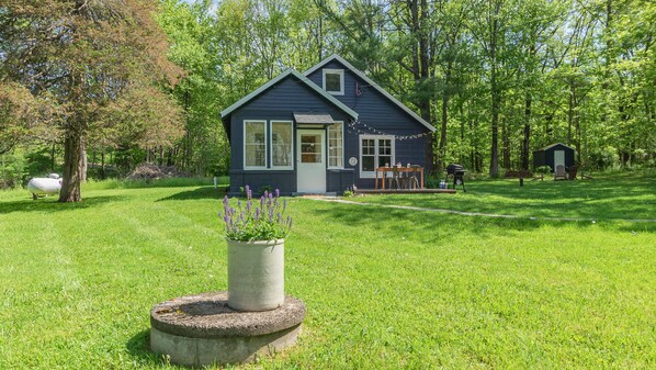 A small, dark blue house with a front porch, surrounded by a lush green lawn and trees. - Whimsical Woodland Cottage in Stone Ridge NY (Stone Ridge)