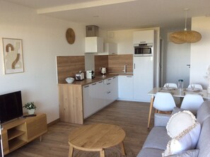 A modern kitchen with white cabinets, a wooden countertop, and a built-in oven. There is a dining area with a table and chairs, and a living room with a sofa and a TV.