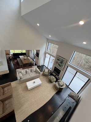 A spacious living area with a high ceiling, large windows, and a fireplace.