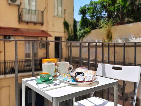A balcony with a table set for breakfast, including a green cup, a white mug, and a plate with food and a drink.