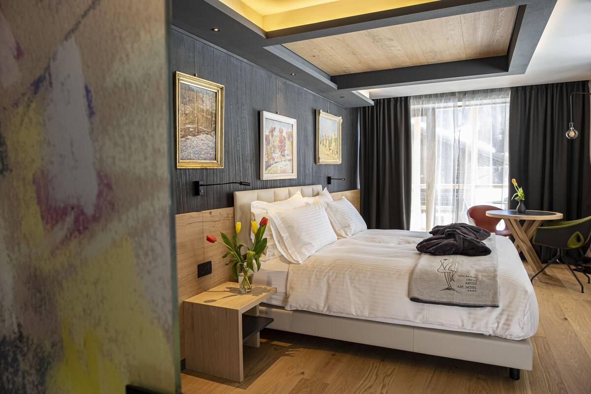 A modern hotel room with a large bed, a bedside table with a vase of flowers, and artwork on the wall.